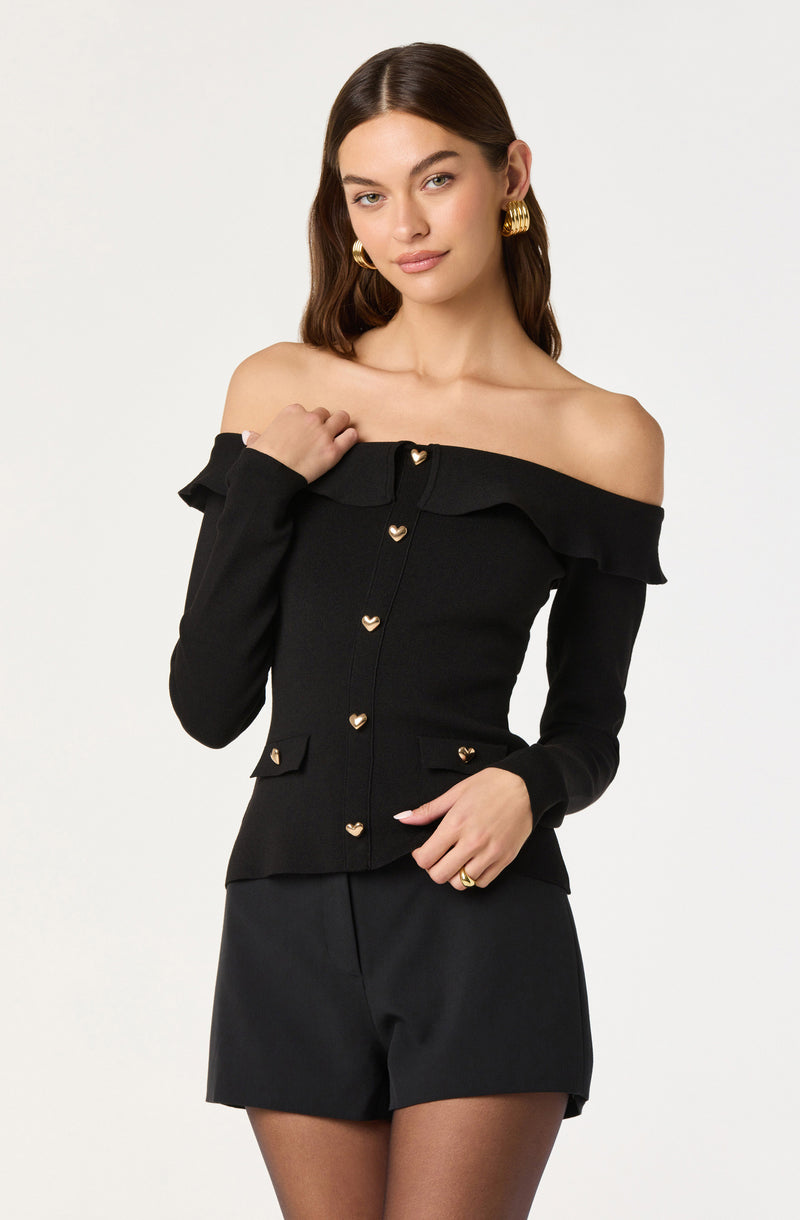 A woman is wearing an off-the-shoulder black top with ruffled detailing along the neckline and long sleeves. The top features gold heart-shaped buttons down the front and on the two small decorative pockets at the waist. She is also wearing black shorts with a tailored fit. She accessorized with large gold hoop earrings and a gold ring on her right hand.