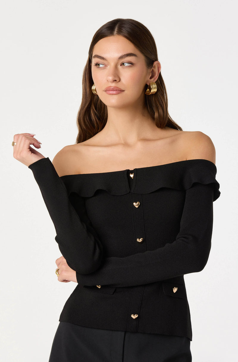 A woman is wearing an off-the-shoulder black sweater with a ruffled neckline and gold heart-shaped buttons down the front. She is accessorized with large gold hoop earrings and a gold ring on her right hand.