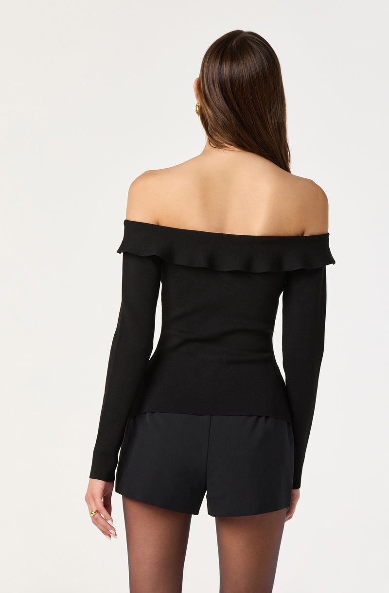 A woman is wearing an off-the-shoulder black top with a ruffled neckline and long sleeves. She pairs it with black shorts, and she has long, straight brown hair.