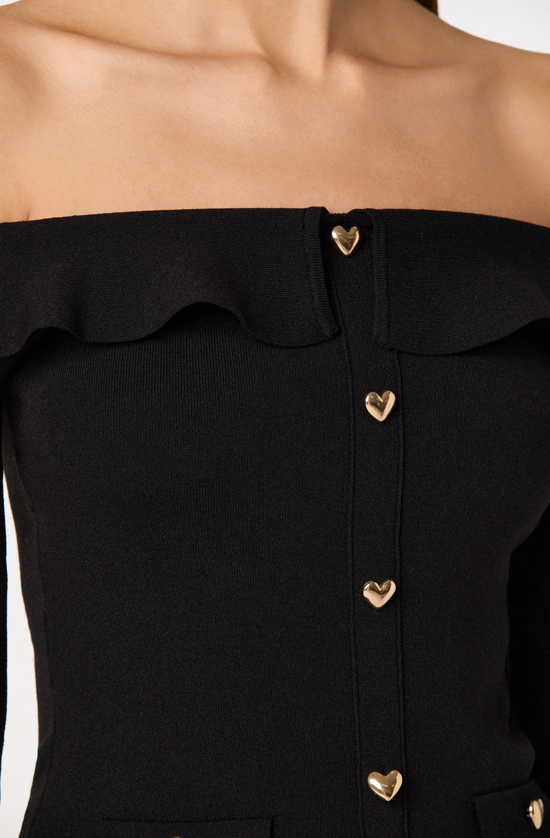 A close-up of a black off-the-shoulder top with a ruffled neckline. The top features a vertical row of gold heart-shaped buttons down the front.