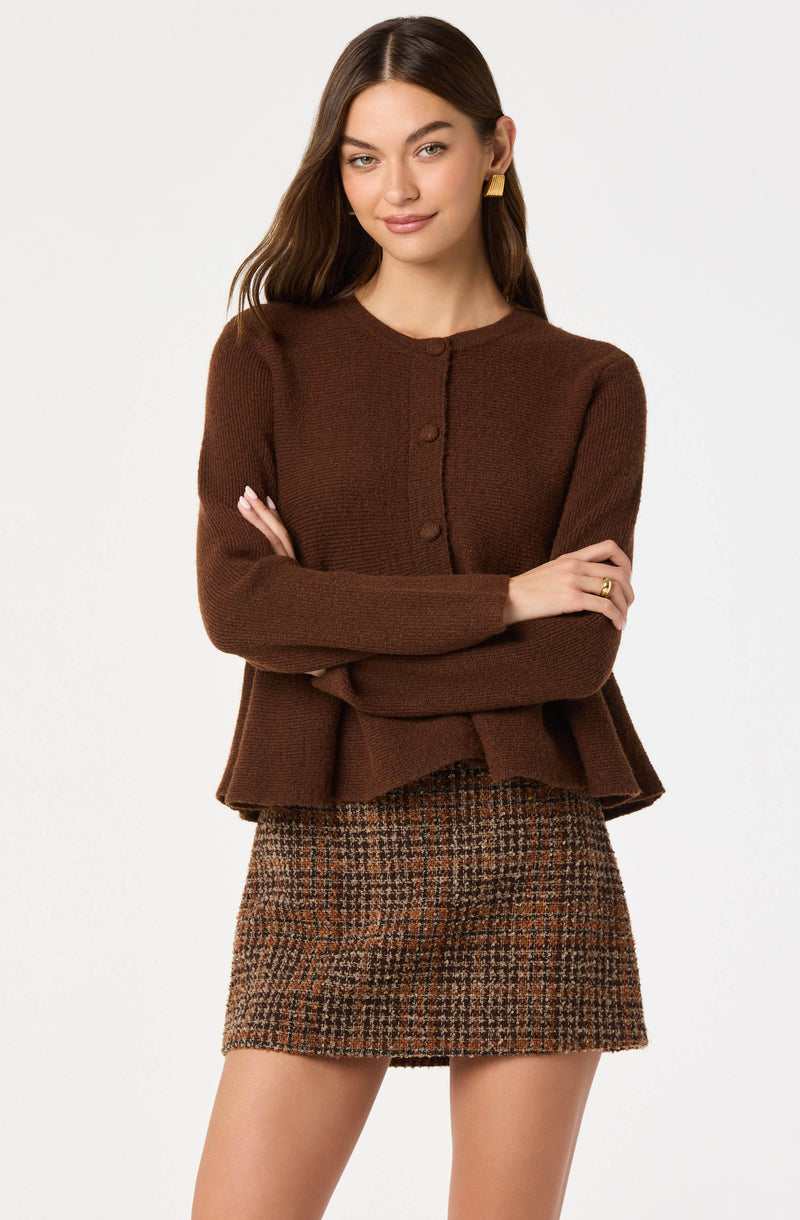 A woman is wearing a brown, long-sleeved, button-up cardigan with a rounded neckline. She has paired it with a high-waisted, plaid mini skirt in shades of brown, orange, and black.