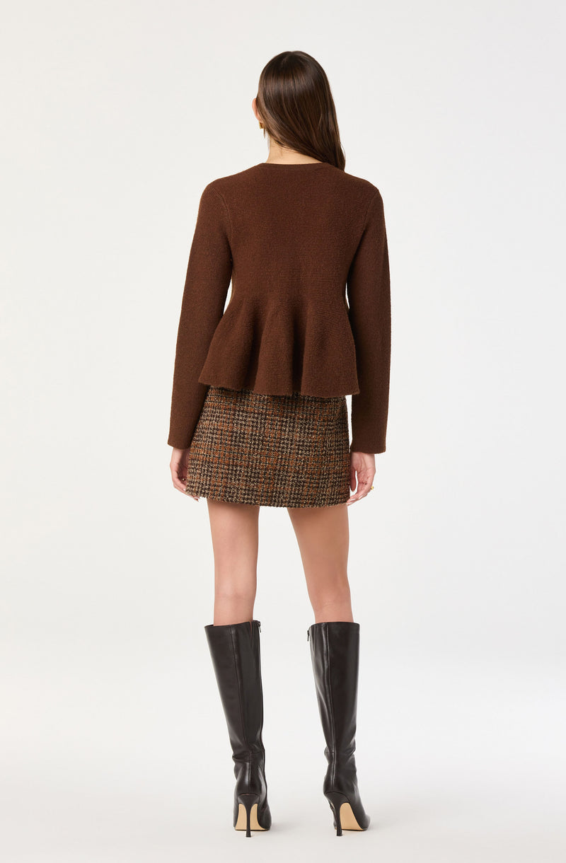 A woman is wearing a brown, long-sleeved, fitted sweater with a peplum detail that flares out at the waist. She is also wearing a high-waisted, plaid mini skirt in shades of brown, orange, and black. She has on tall black leather knee-high boots with pointed toes and stiletto heels.