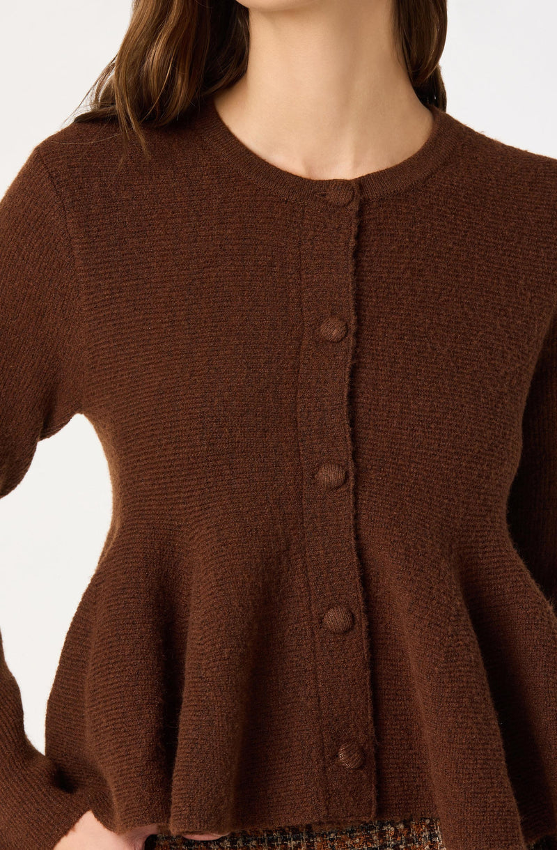A woman is wearing a long-sleeved, brown knitted cardigan with a round neckline. The cardigan features a row of buttons down the front and has a slightly flared hem. The fabric appears textured, giving it a cozy look.