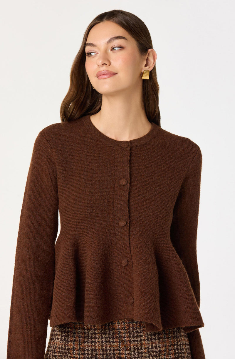 A woman is wearing a brown, textured knit cardigan with a round neckline, buttoned down the front with matching brown buttons. The cardigan has a slightly flared, peplum-style hem. She is also wearing plaid patterned pants with shades of brown.