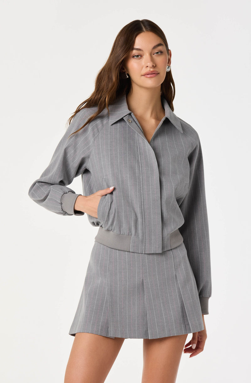 A woman is wearing a gray, pinstriped, collared shirt with long sleeves and a matching gray, pinstriped, pleated mini skirt. The shirt has a cropped style with a ribbed waistband, and the skirt has a high waist and a small slit in the front.
