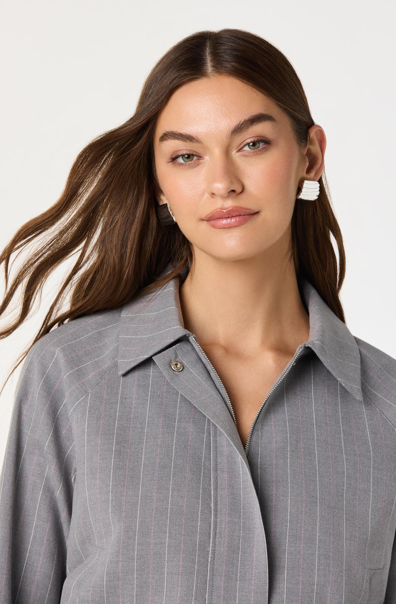 A woman is wearing a gray pinstripe jacket with a collar and a partially zipped front. The jacket has subtle thin vertical pink stripes. She is also wearing large white earrings.