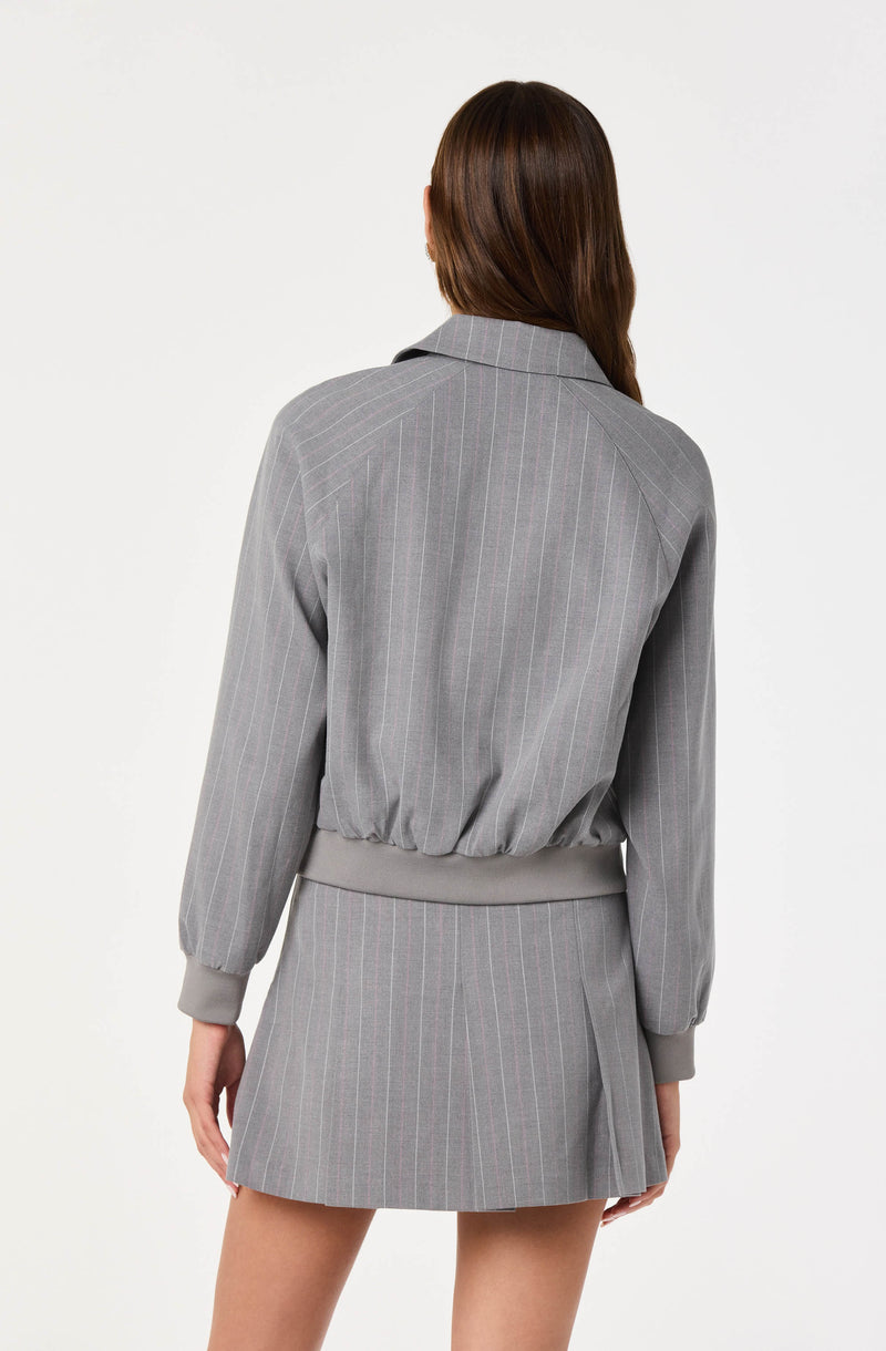 A person is wearing a matching gray pinstripe jacket and skirt. The jacket has long sleeves with ribbed cuffs and a ribbed waistband, and a classic collar. The skirt is a high-waisted, above-the-knee length with pleats on the side. The fabric has a subtle vertical stripe pattern. The person's hair is long and brown, and they are standing with their back to the camera against a plain white background.