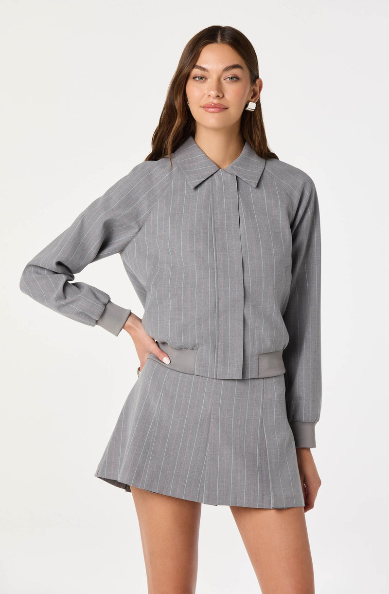 A woman is wearing a gray set consisting of a long-sleeved, collared jacket with vertical pinstripes and matching pleated mini skirt, both made of a tailored fabric. The jacket has cuffs and a front closure with pleats at the neckline. She has long, wavy brown hair and is accessorized with large, white geometric earrings.