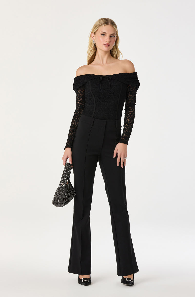 A woman is wearing a black off-the-shoulder top with long, sheer lace sleeves and a textured bodice that ties at the neckline. She pairs the top with high-waisted, tailored black trousers that have a slight flare at the bottom. She is also holding a small, black handbag decorated with rhinestones, and is wearing black and white pointed-toe shoes with a buckle detail.