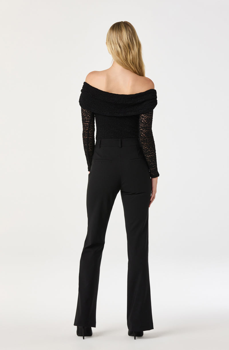 A woman is wearing an off-the-shoulder black sweater with a textured, fuzzy fabric. The sweater has long sleeves made of a lace-like, see-through material. She pairs this with high-waisted, straight-leg black pants. The overall look is stylish and elegant, with an emphasis on the black clothing items.