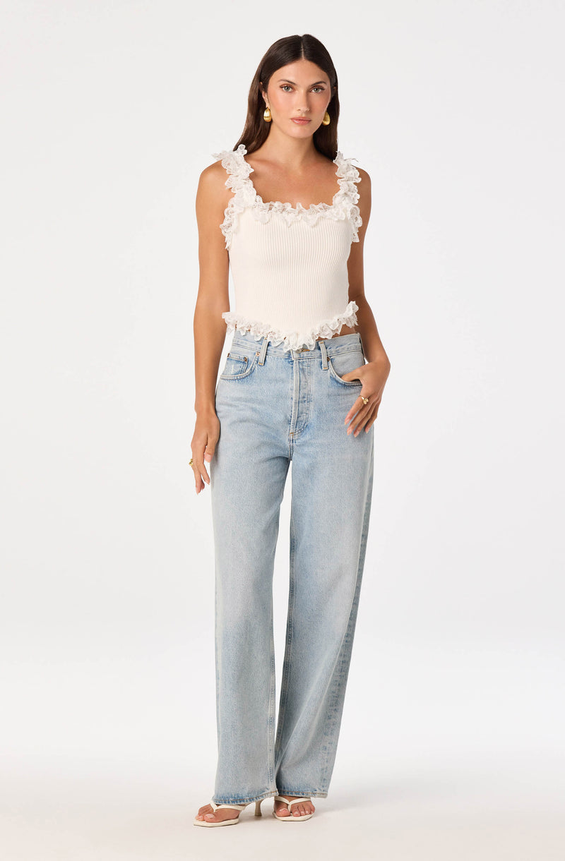Woman wearing a sleeveless white top with ruffled details and light-wash, high-waisted, wide-leg jeans.