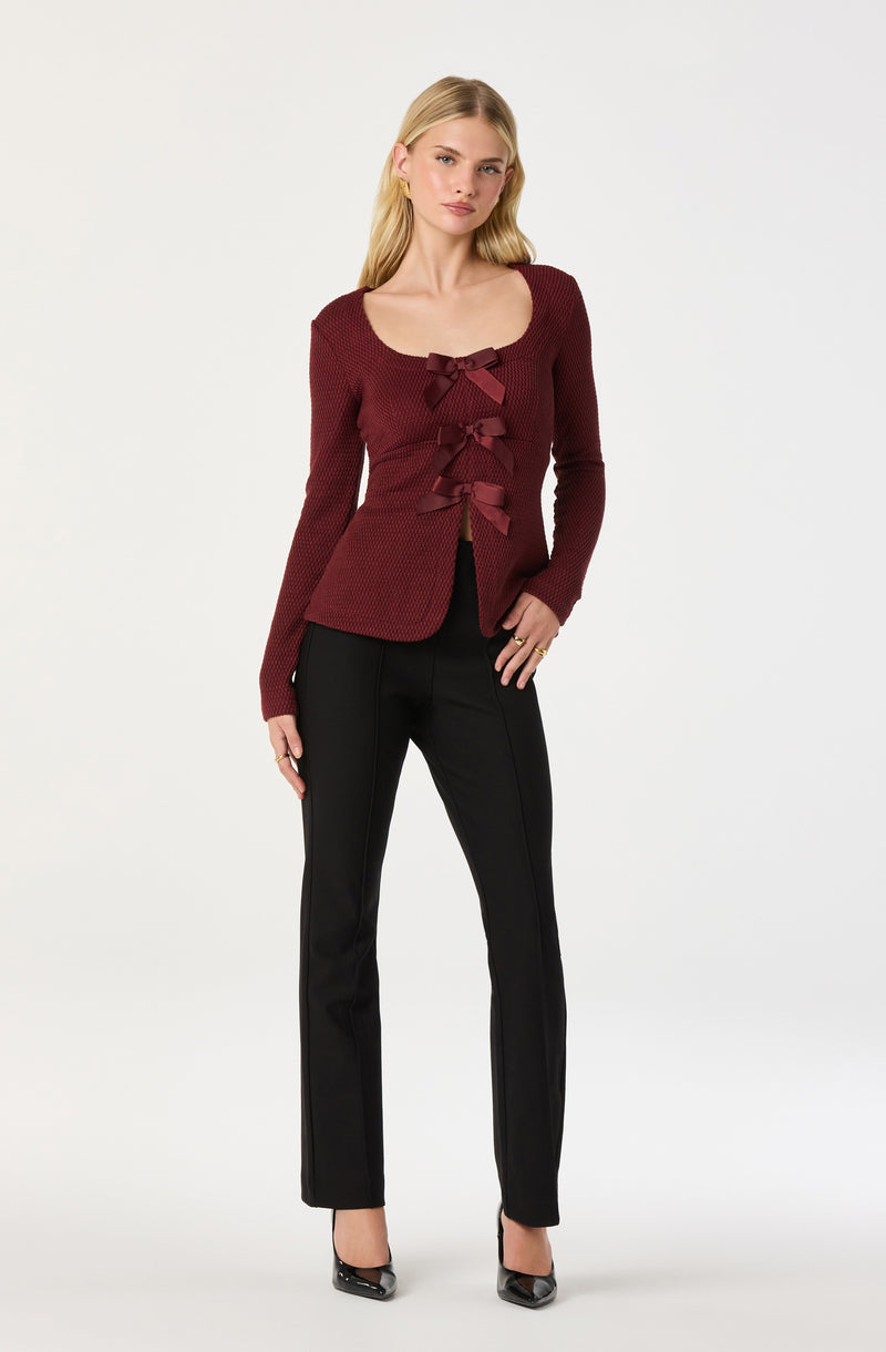 A woman is wearing a long-sleeved, maroon knit cardigan with a scoop neckline and three decorative bows down the front. She pairs it with black, straight-leg trousers and black pointed-toe heels.