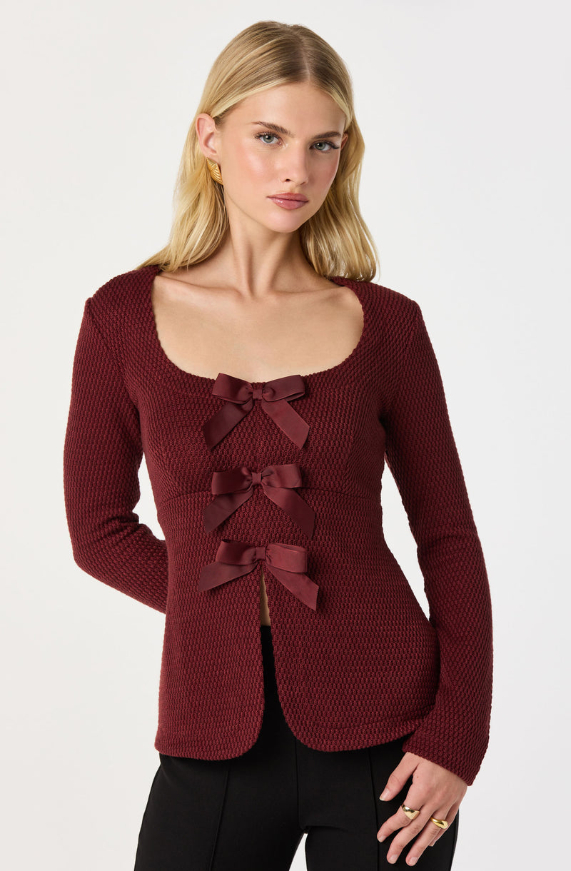 A woman is wearing a fitted, long-sleeved, maroon knit blazer with a rounded neckline. The blazer features three decorative maroon satin bows running vertically down the front.