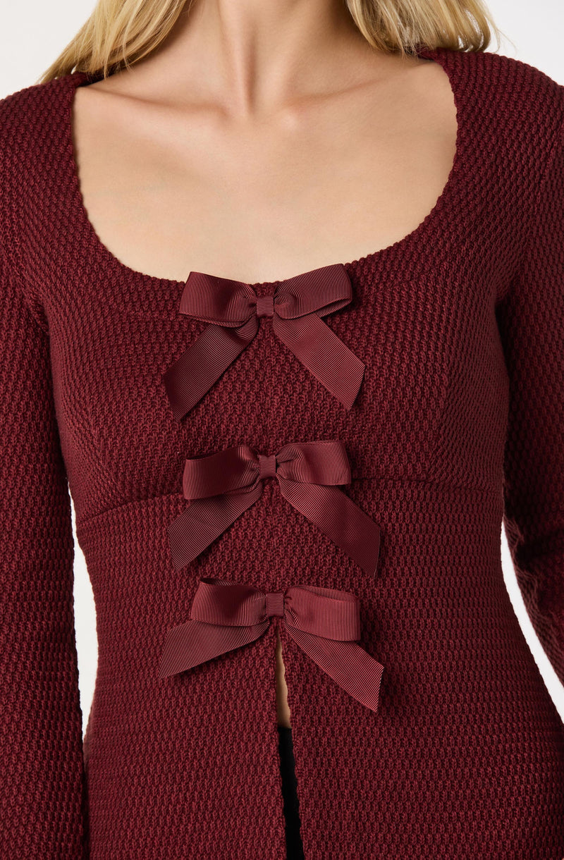 A person is wearing a red textured knit cardigan with a round neckline. The cardigan features three large, matching red satin bows down the front. The fabric has a honeycomb-like pattern, and the cardigan is fitted to the body.