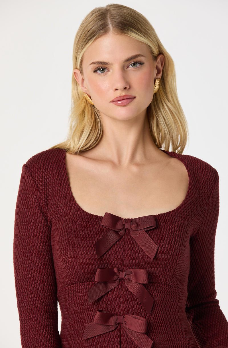 A woman with blonde hair wearing a long-sleeved, textured burgundy dress featuring three satin bows down the front.