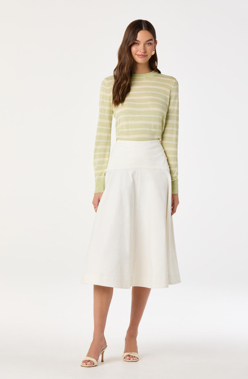 A woman is wearing a light green and white horizontally striped long-sleeve sweater, paired with a high-waisted, white A-line midi skirt that reaches below her knees. She is also wearing cream-colored open-toe high-heeled sandals.