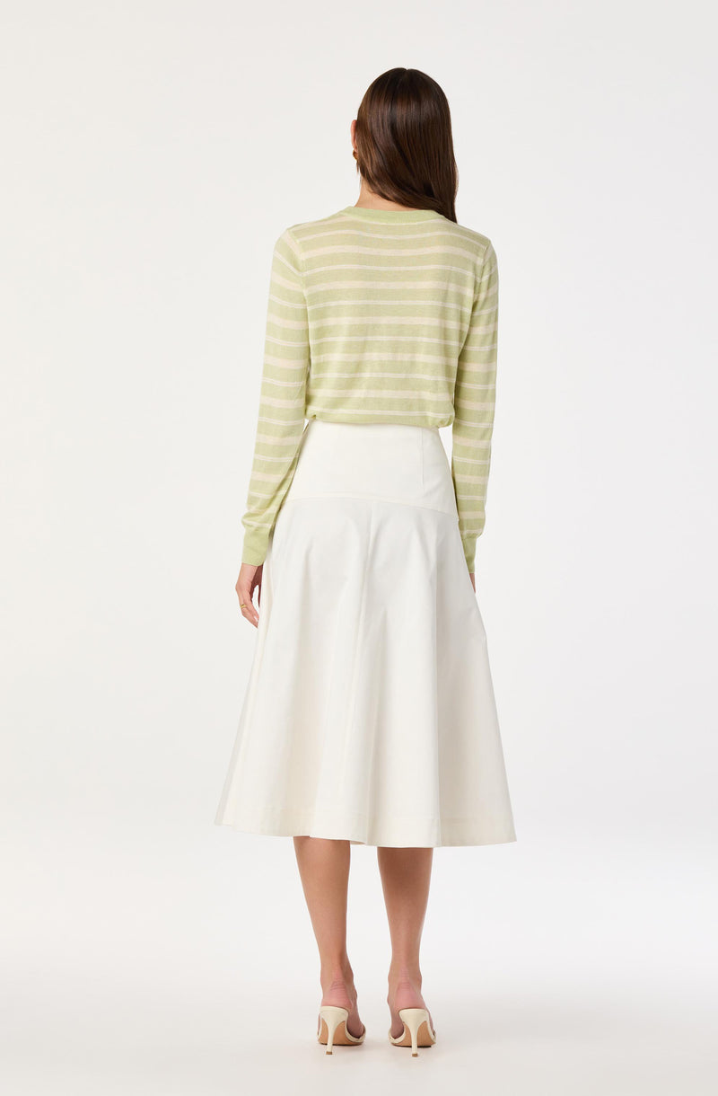 A woman is wearing a light green long-sleeve sweater with horizontal white stripes and a high-waisted, white, A-line midi skirt that falls below the knees. She is also wearing beige high-heeled shoes.