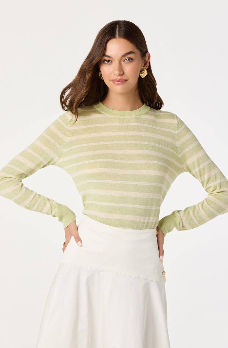 A woman is wearing a light green long-sleeve sweater with horizontal beige stripes and a cream-colored A-line skirt. She has her hands on her hips, and the background is plain white.