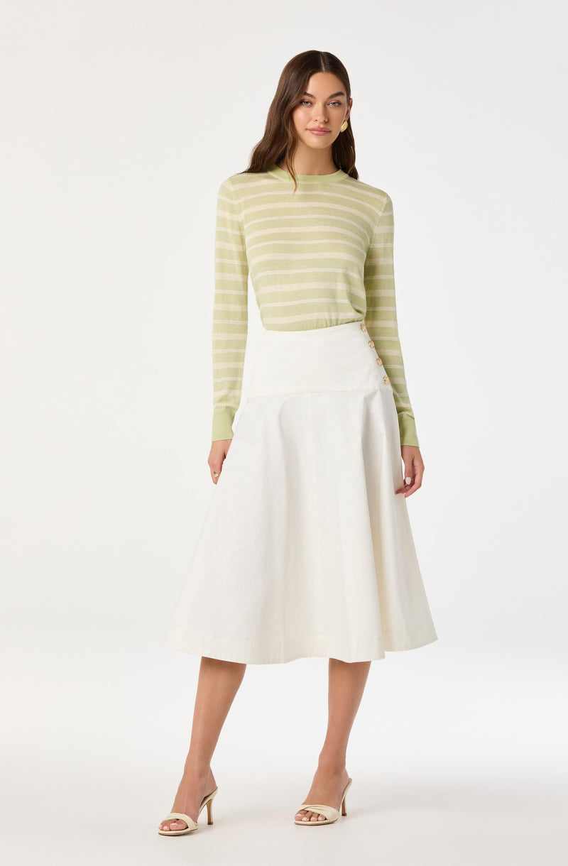 A woman is wearing a long-sleeved, light green and beige horizontally striped sweater tucked into a high-waisted, white, A-line midi skirt with buttons on the side. She is also wearing beige high-heeled sandals.