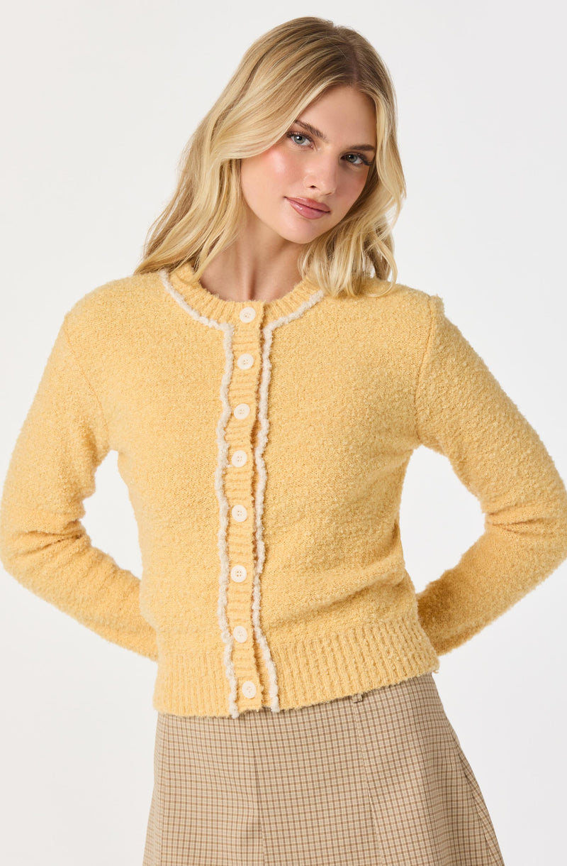 A woman is wearing a yellow, textured knit cardigan with white piping along the edges and around the round neckline. The cardigan has a row of white buttons down the front. She is also wearing a beige skirt with a small checkered pattern.