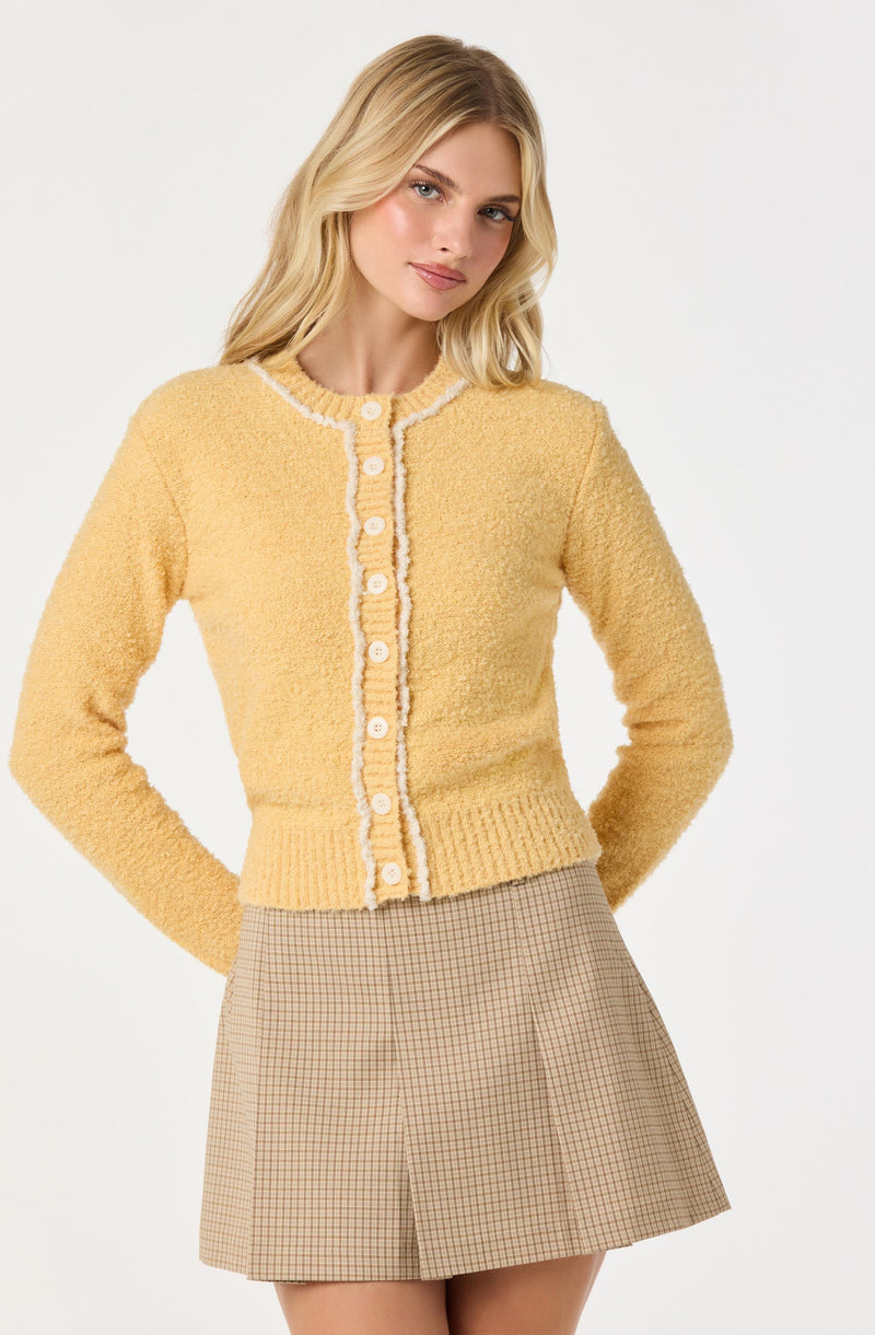 A woman is wearing a yellow, textured knit cardigan with white trim along the neckline, front opening, and hem. The cardigan has white buttons down the front. She pairs it with a beige, plaid A-line skirt.