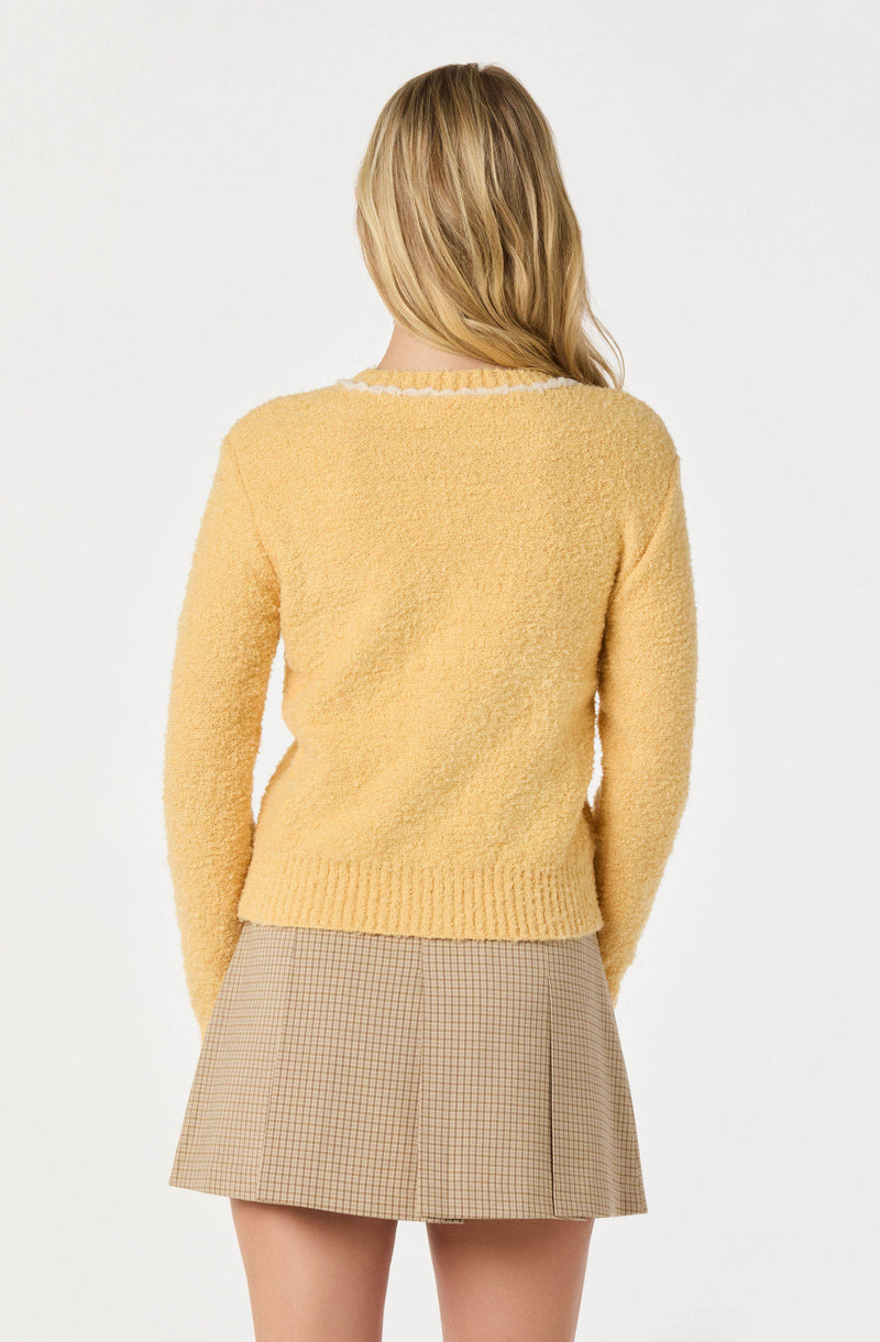 A person is wearing a soft, fluffy, yellow sweater with a slightly textured appearance. The sweater has a round neckline, which is accented with a contrasting white trim. They are also wearing a beige plaid skirt that reaches mid-thigh, featuring a subtle checkered pattern in shades of brown and black.