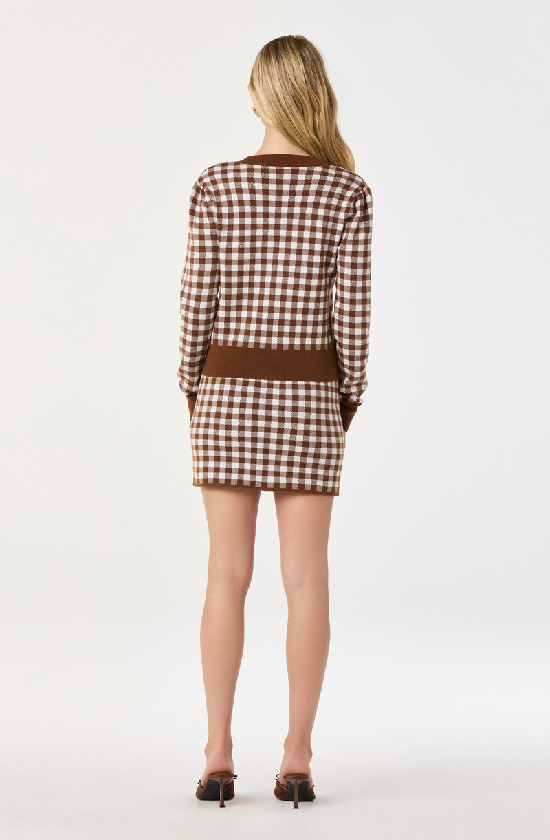 A woman is wearing a long-sleeved, brown and white checkered dress with a solid brown band at the horizon. The dress has a fitted style, and she is wearing brown high-heeled shoes. She is standing with her back to the camera, with her arms relaxed at her sides.