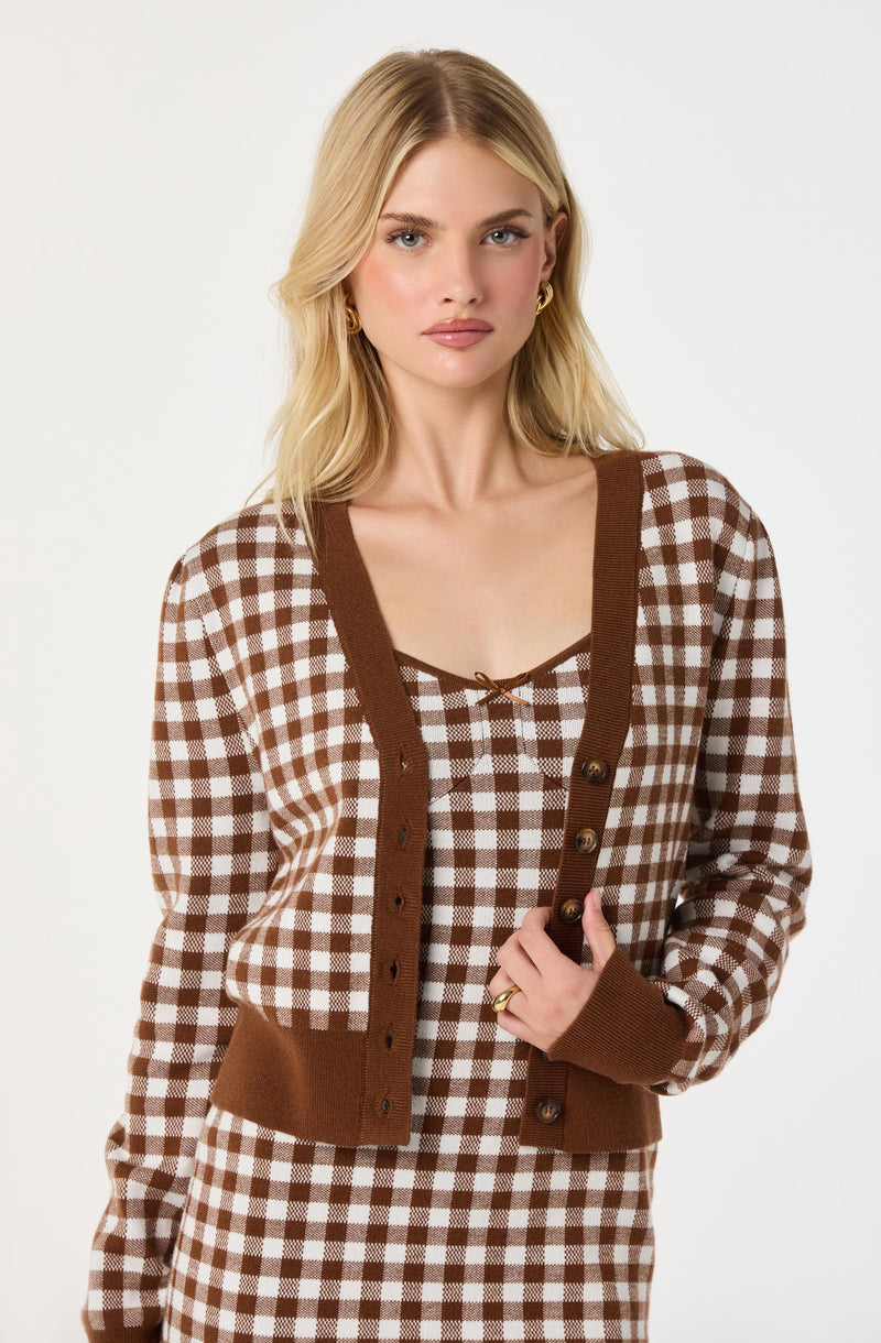A woman is wearing a brown and white checkered cardigan with brown trim along the neckline, button placket, and cuffs. Underneath, she has on a matching checkered top with a small bow detail at the neckline.