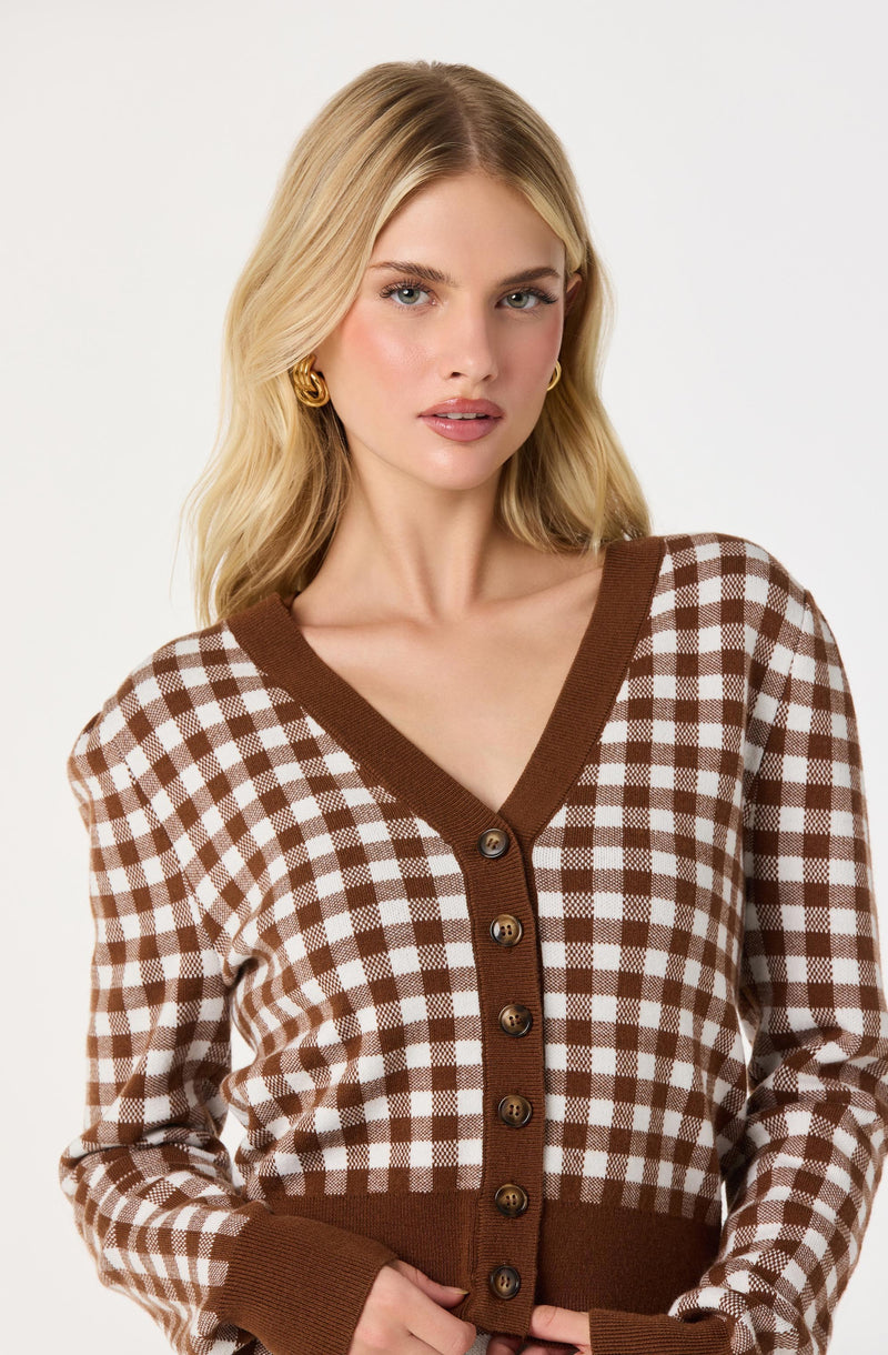 A woman wearing a brown and white checkered cardigan with a V-neckline, brown trim along the neckline and front opening, and brown buttons down the center. The cardigan has long sleeves with brown ribbed cuffs.