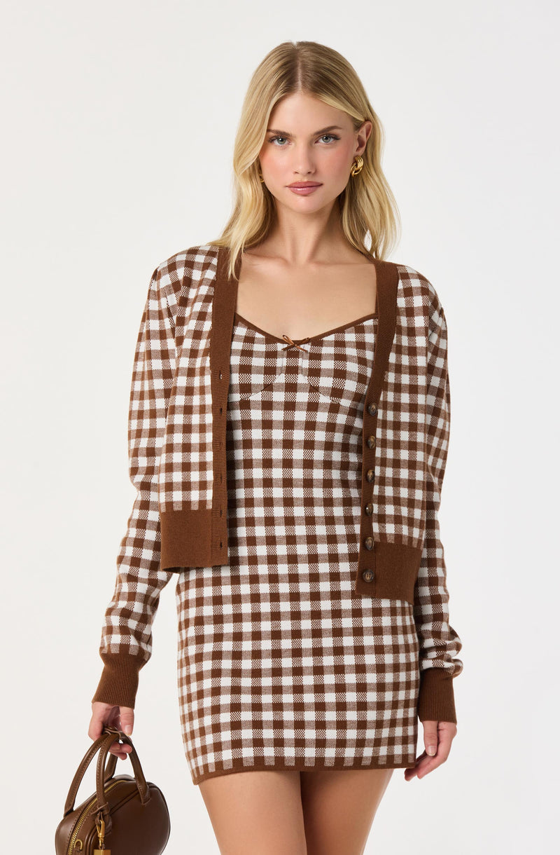A woman is wearing a brown and white checkered dress with a matching brown cardigan that has buttons down the front and ribbed cuffs. The dress has thin straps and a small bow at the neckline. She is holding a small brown handbag in her left hand.