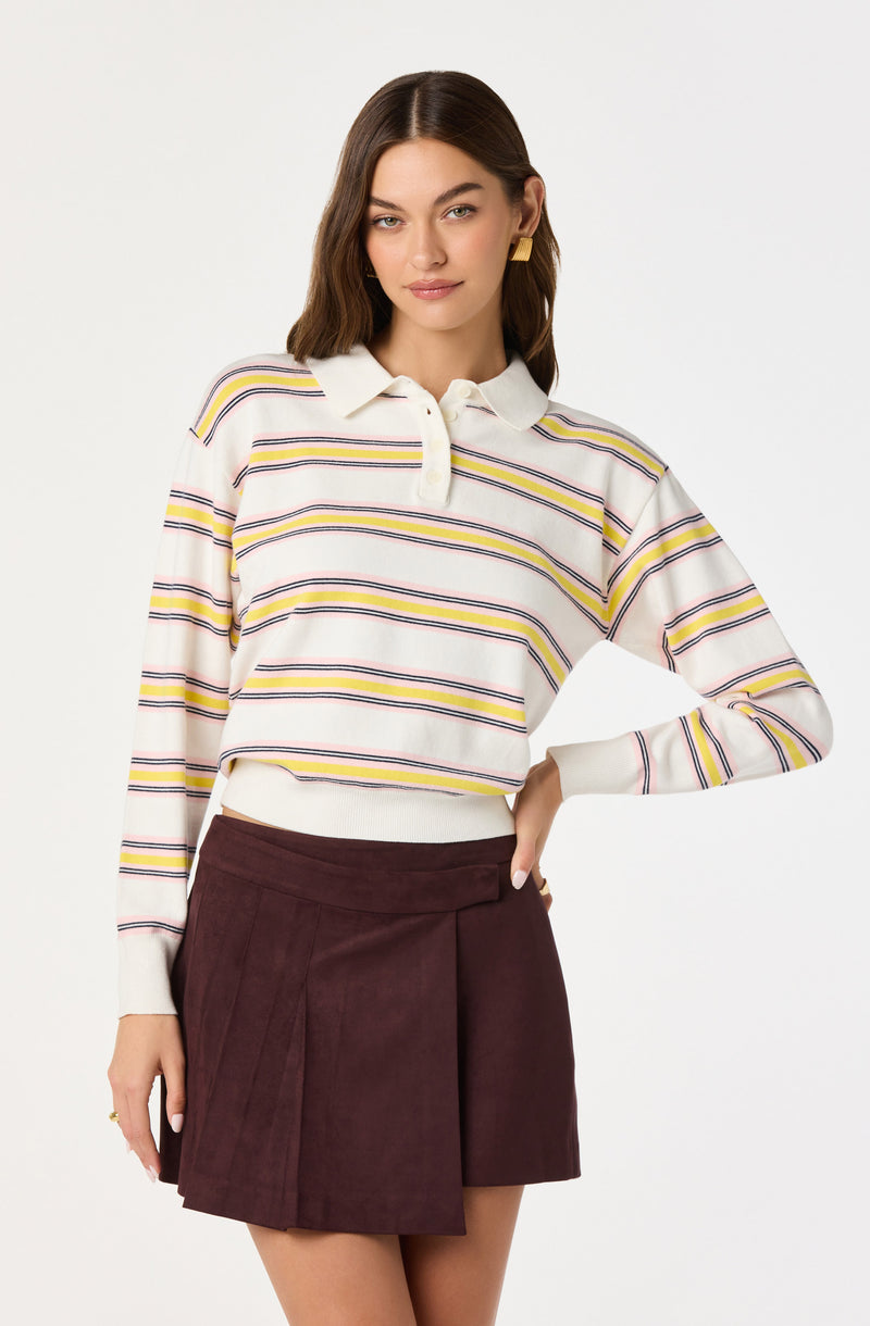 A woman is wearing a long-sleeved, collared polo sweater with horizontal stripes in yellow, purple, pink, and navy on a white background. She pairs it with a dark burgundy, high-waisted mini skirt that features pleats on one side. She has shoulder-length brown hair, gold square earrings, and a subtle smile.