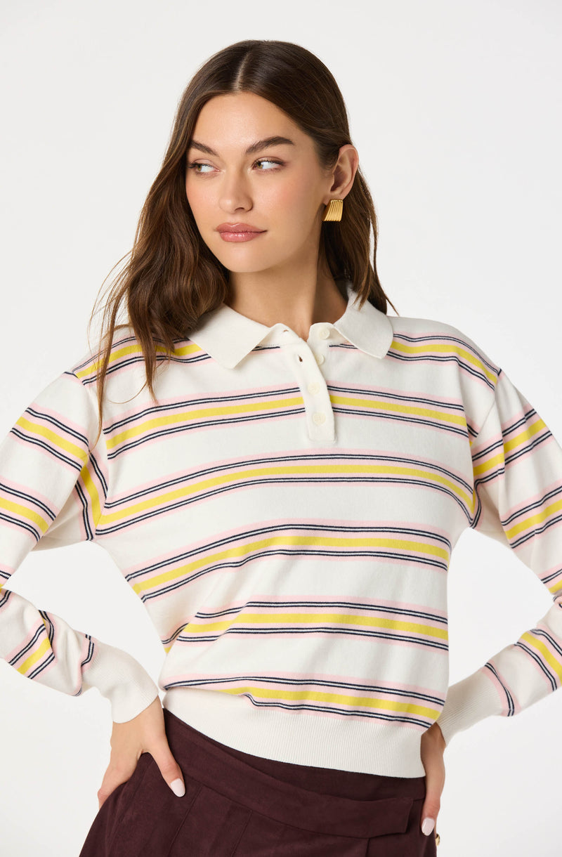 A woman is wearing a long-sleeved, collared sweater with horizontal stripes in yellow, pink, black, and white. The sweater has a buttoned placket at the collar and ribbed cuffs at the wrists.