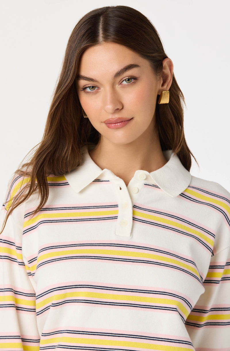 A woman is wearing a long-sleeved, collared shirt with horizontal stripes in yellow, pink, black, and white. The shirt has a white collar and a button-up placket. She is also wearing gold, geometric-shaped earrings.