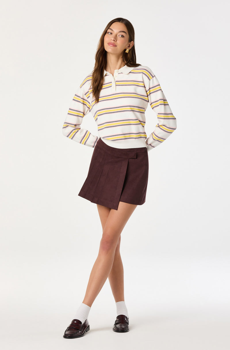 A woman is wearing a long-sleeved, collared shirt with horizontal stripes in white, yellow, and purple. She pairs it with a high-waisted, dark burgundy skirt that has a front pleat. She completes the outfit with white socks and dark loafers.