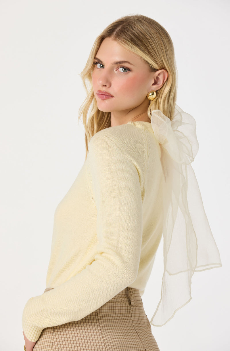 A woman is wearing a light yellow, long-sleeved sweater with a sheer, ruffled bow detail on the shoulder, paired with a beige plaid skirt. She has blonde hair, and is accessorized with large, gold, hoop earrings.