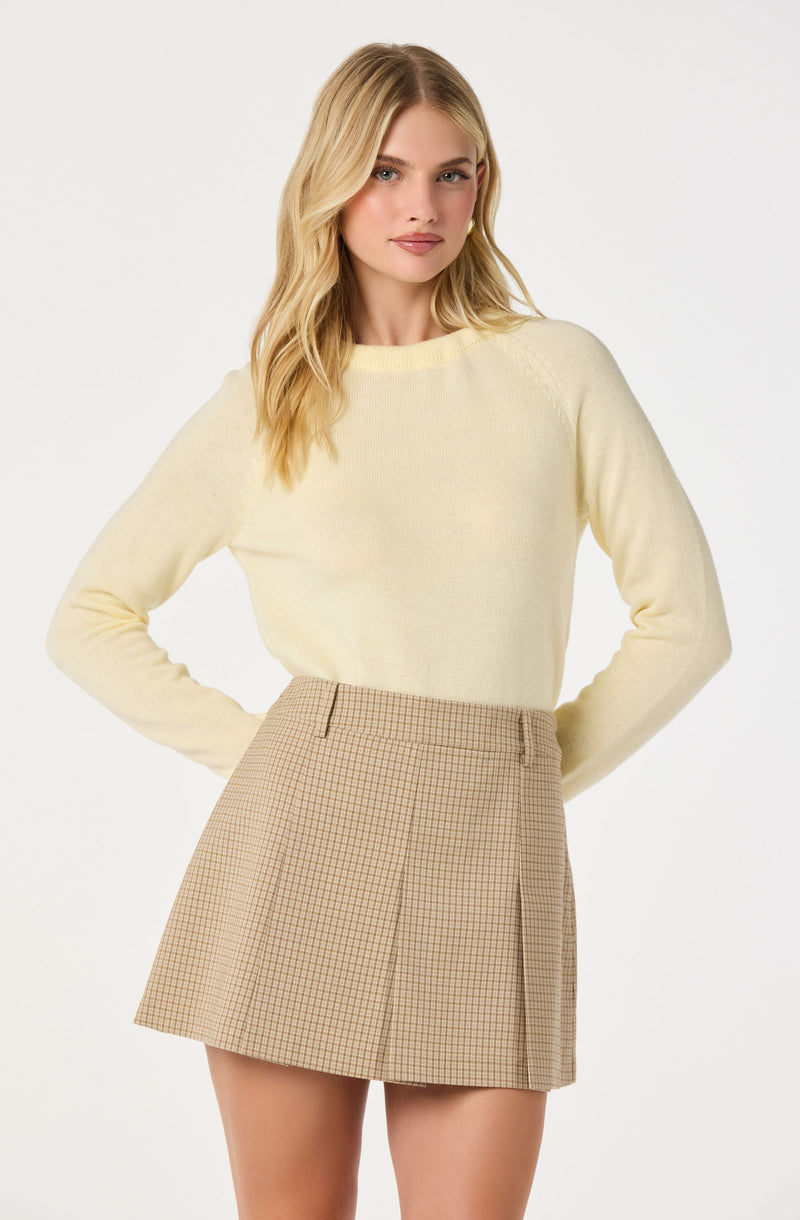 A young woman is wearing a light yellow long-sleeve sweater with a round neckline and beige high-waisted skirt featuring a small checkered pattern and pleats on the front. The outfit is simple and stylish, with a soft and neutral color palette.