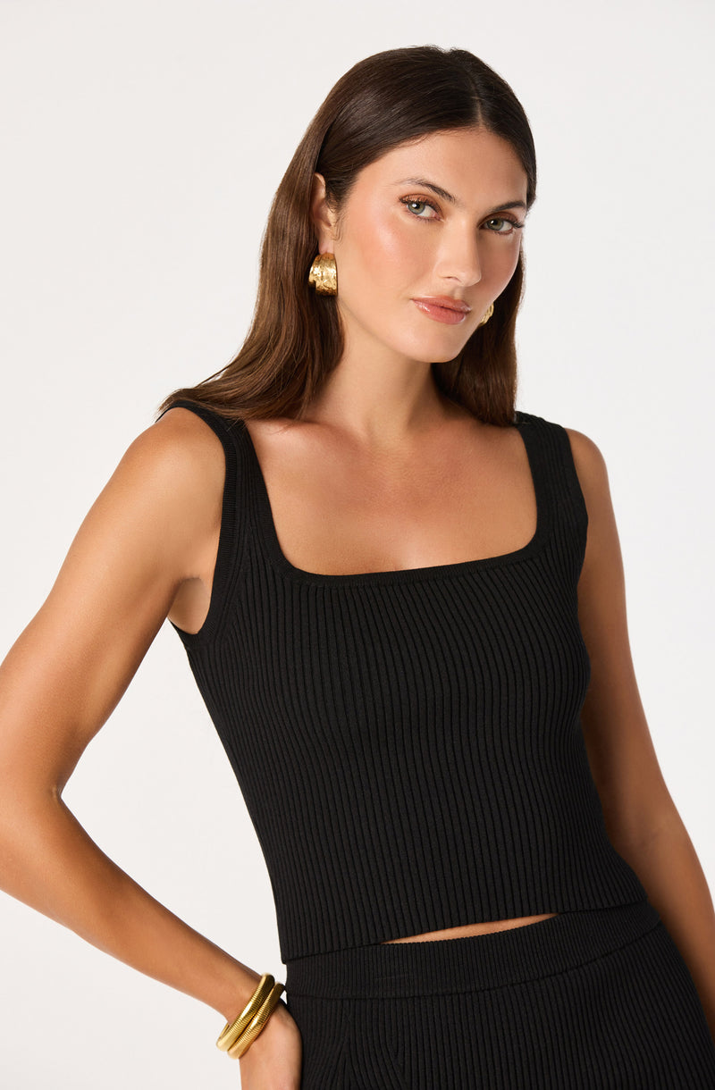 Woman wearing a black ribbed tank top and matching black pants, accessorized with gold earrings and bracelets.