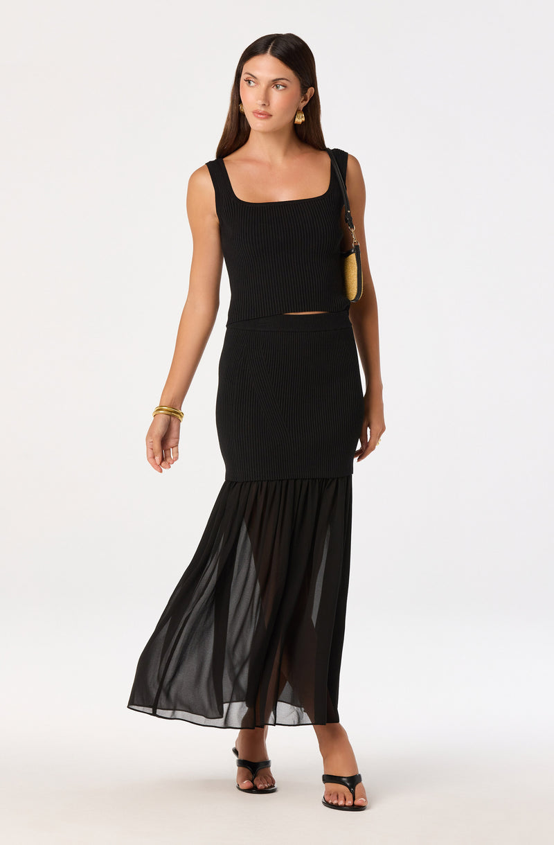 Woman in a black ribbed sleeveless top and matching high-waisted skirt with sheer fabric at the bottom.