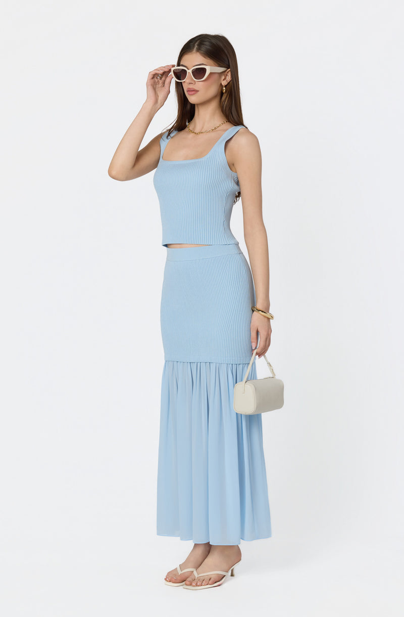 Woman wearing a light blue sleeveless crop top and matching long skirt with ruffle detail, accessorized with white sunglasses and beige sandals.