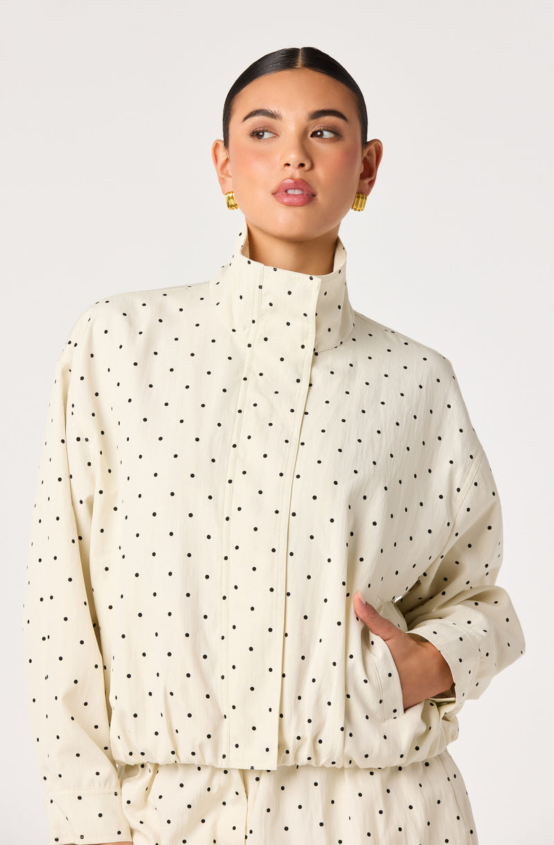 Woman in a cream high-collared polka dot jacket with gold earrings.