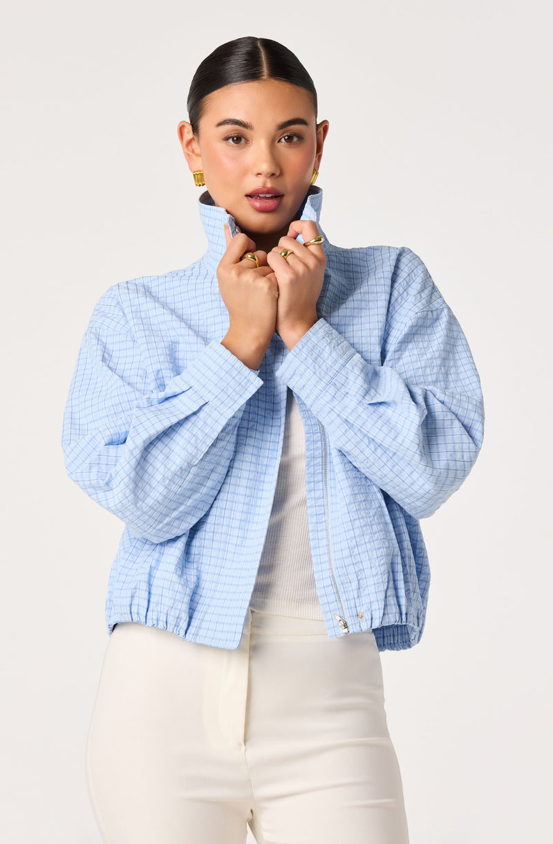 Woman wearing a light blue checkered zip-up jacket over a white top, with white pants.