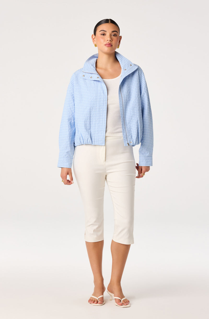Woman wearing a light blue checkered jacket with a hood, white top, cream cropped pants, and white sandals.