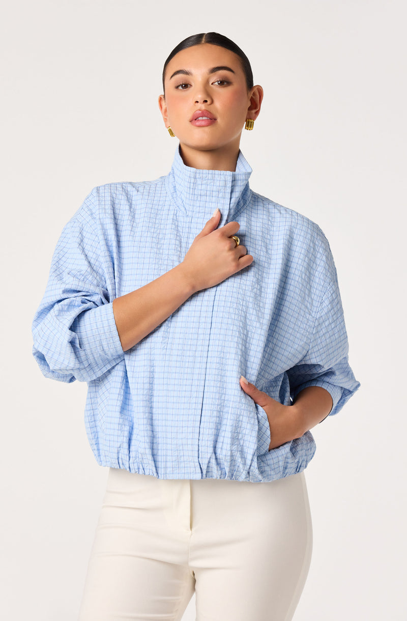 Woman wearing a light blue checkered high-neck jacket and white pants.
