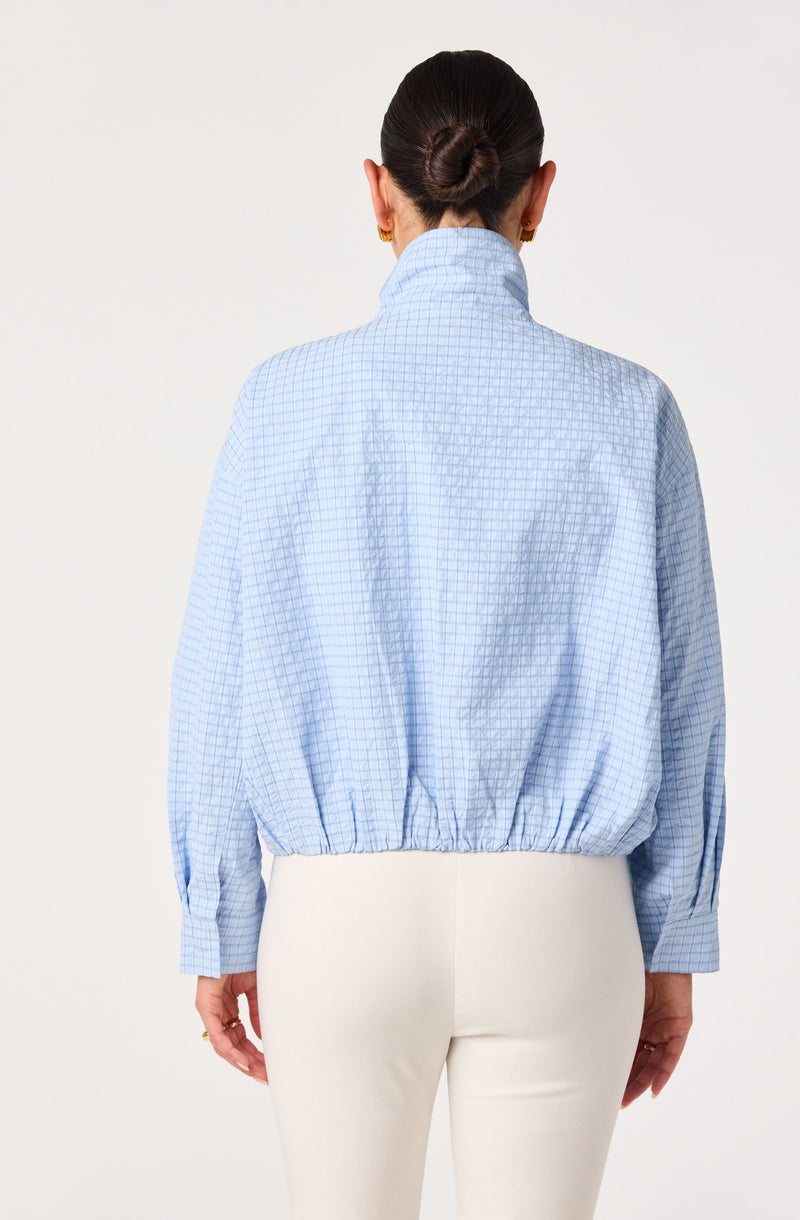 Back view of a woman wearing a light blue checkered jacket with a high collar and gathered hem, paired with white pants.