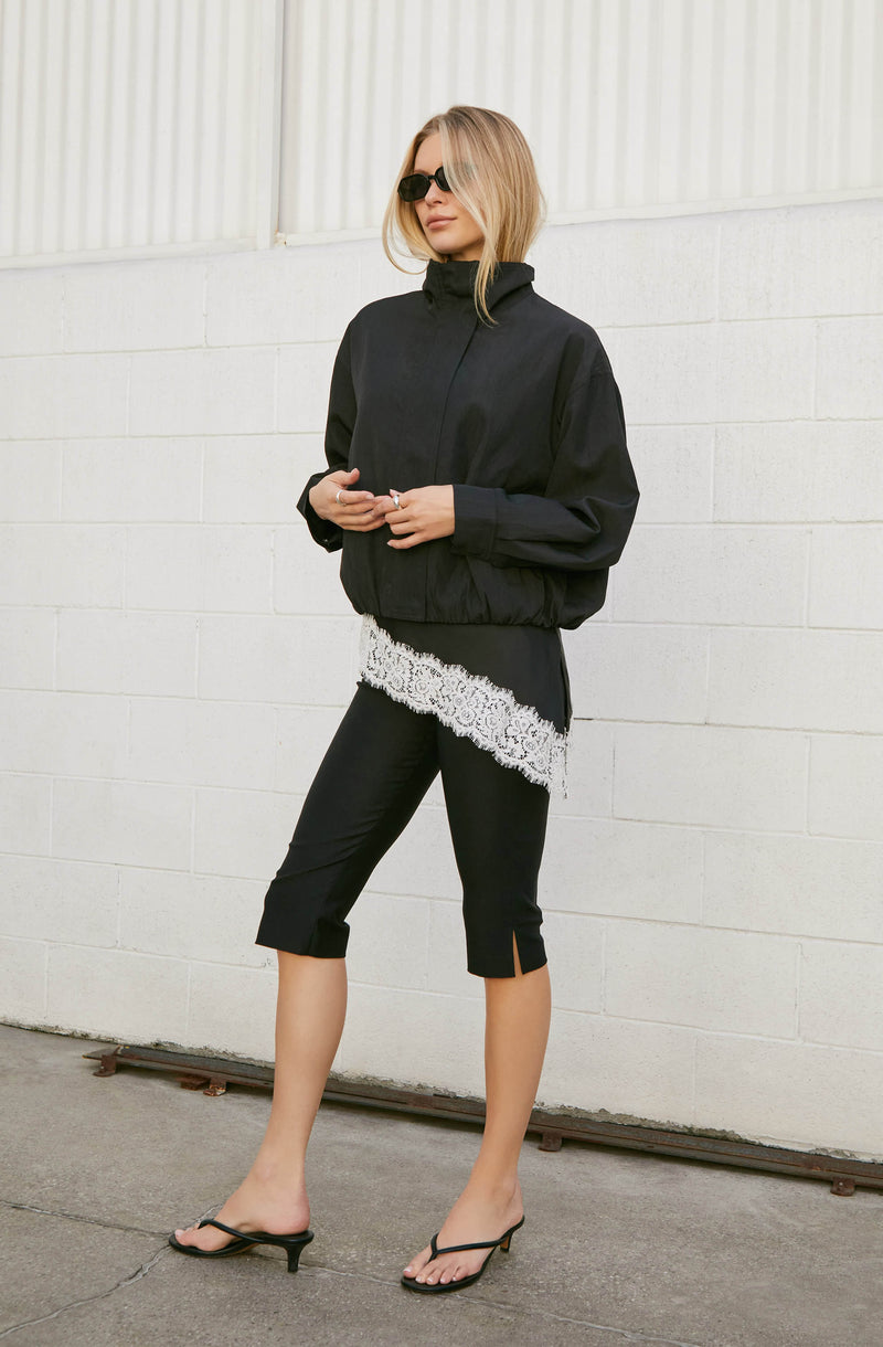 Woman wearing a black zip-up jacket, lace-trimmed top, black capri pants, and black heels.