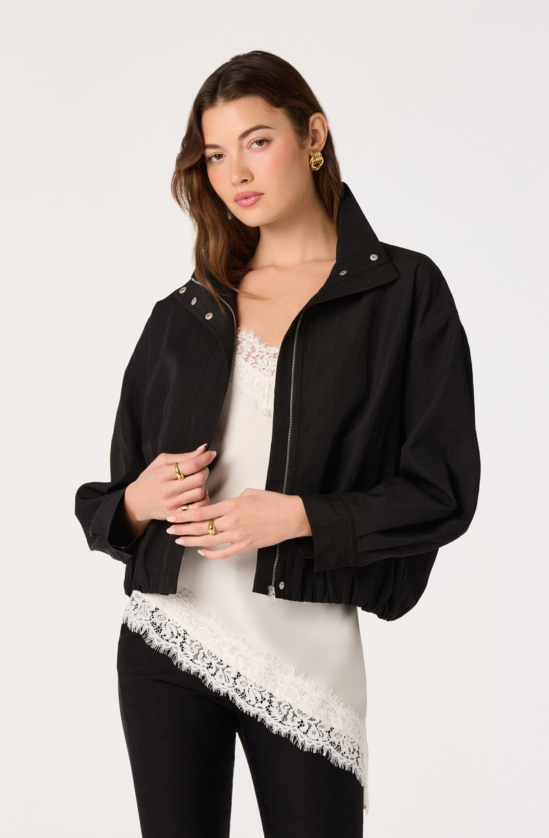 Woman wearing a black jacket over a white lace-trimmed top with black pants.