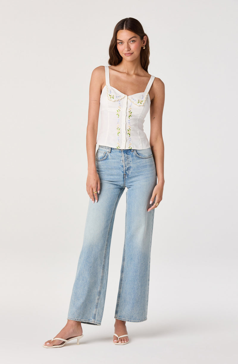 Woman in a white, sleeveless, embroidered top with ruffle strap details and light blue wide-leg jeans.