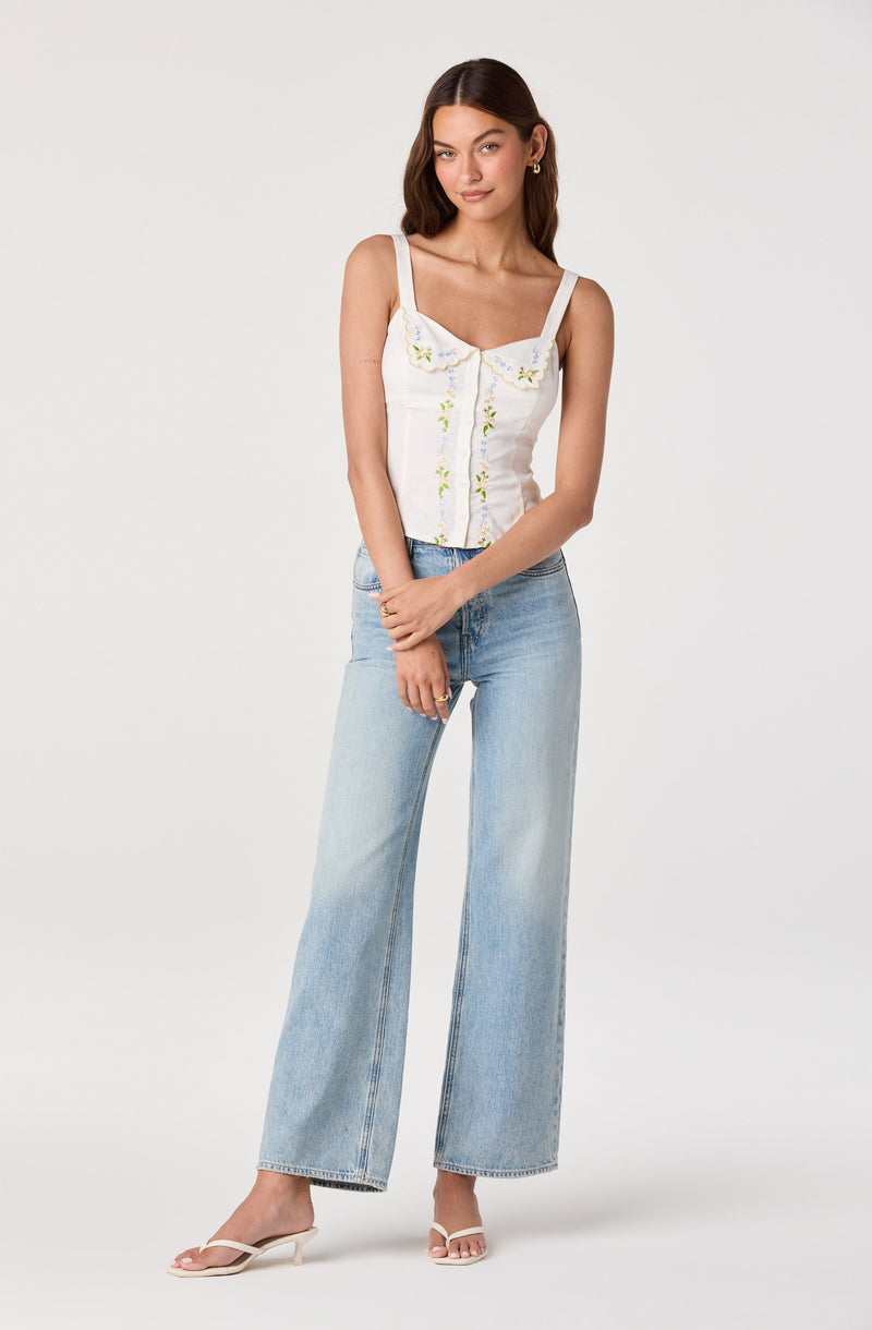 Woman wearing a white sleeveless top with embroidered details and light blue wide-leg jeans.