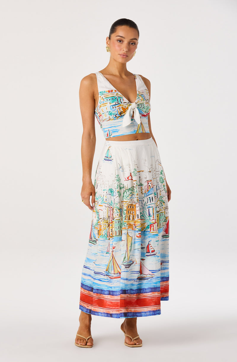 Woman wearing a white sleeveless crop top and a long skirt with a colorful seaside village and sailboats print.
