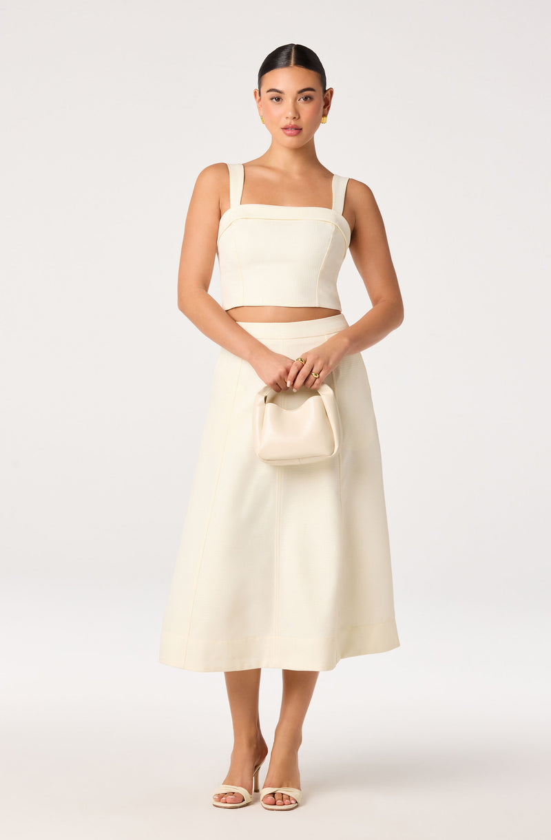 Woman wearing a cream crop top with wide straps and a matching high-waisted, A-line skirt, holding a small clutch.